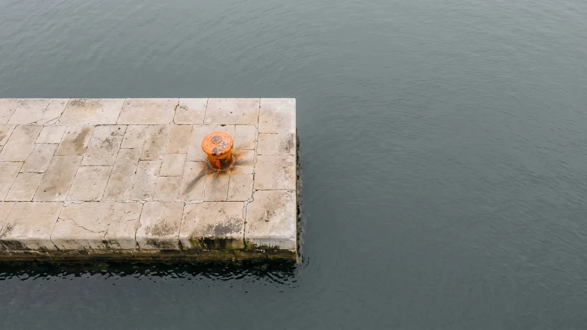 El extremo de un espigón de piedra sobre el mar gris, visto desde arriba, con un noray naranja.